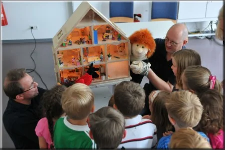 Feuerwehrmann erklärt einer Gruppe von Kindern anhand eines Modells und mit einer Puppe die Risiken des Feuers
